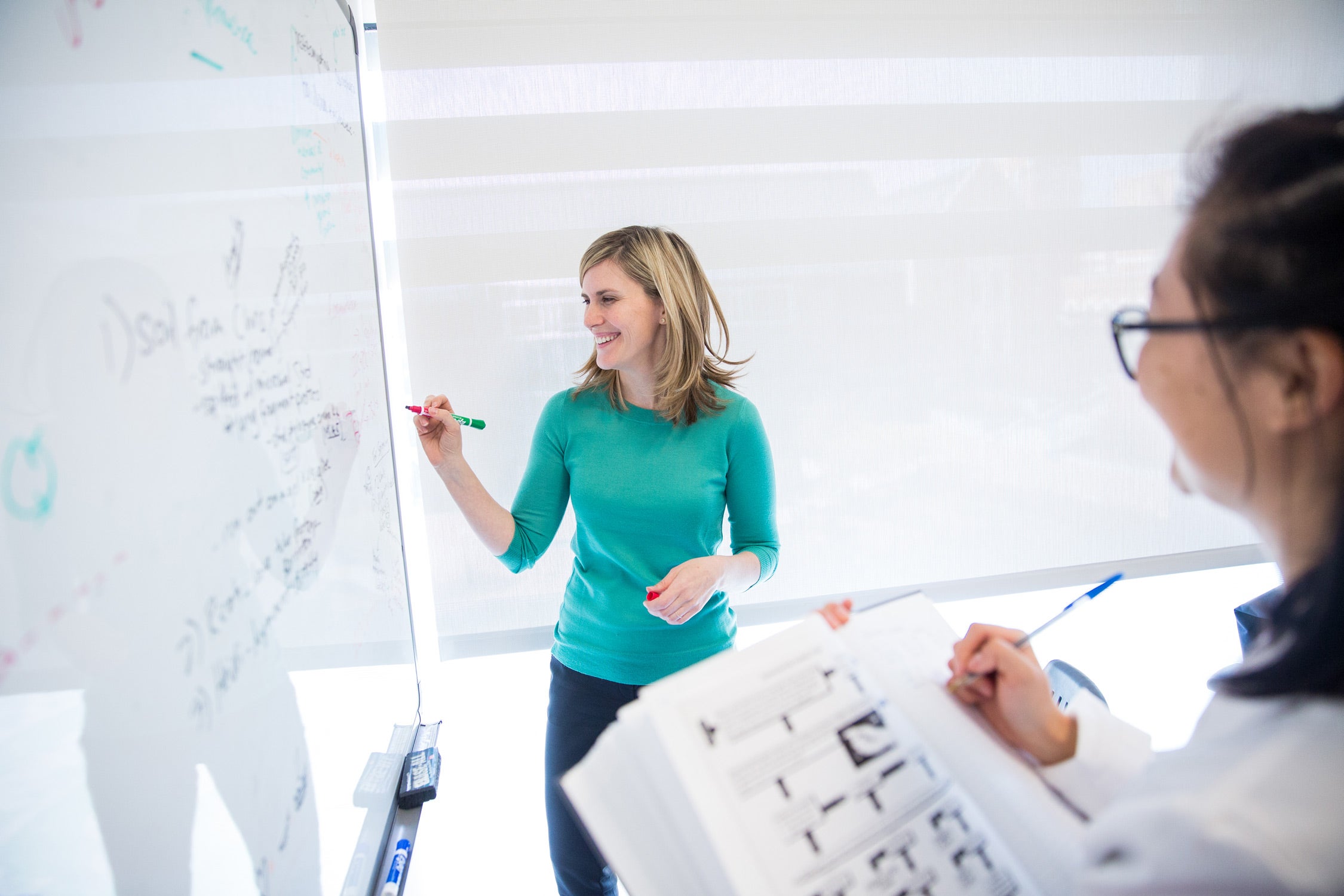 faculty member writing on white board, smiling. While student takes not in textbook.