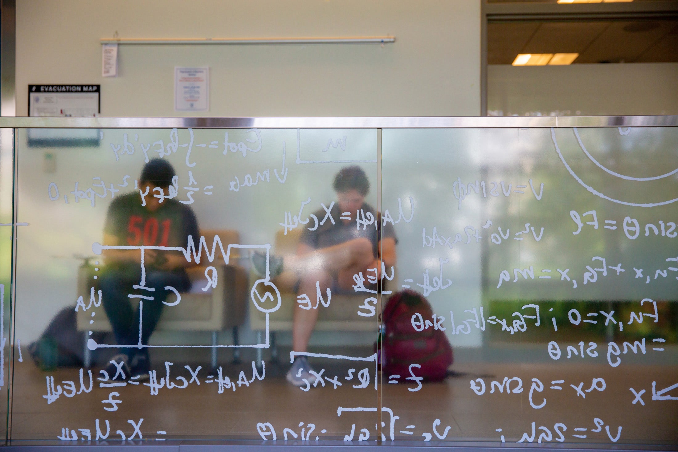 equations written on a clear board. Behind two students out of focus sitting down.
